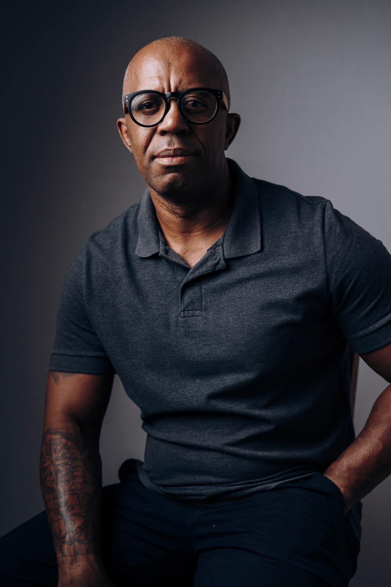 Retrato editorial de homem de polo cinza com óculos e tatuagem no antebraço, fundo cinza, fotografado por Lucian Rabello