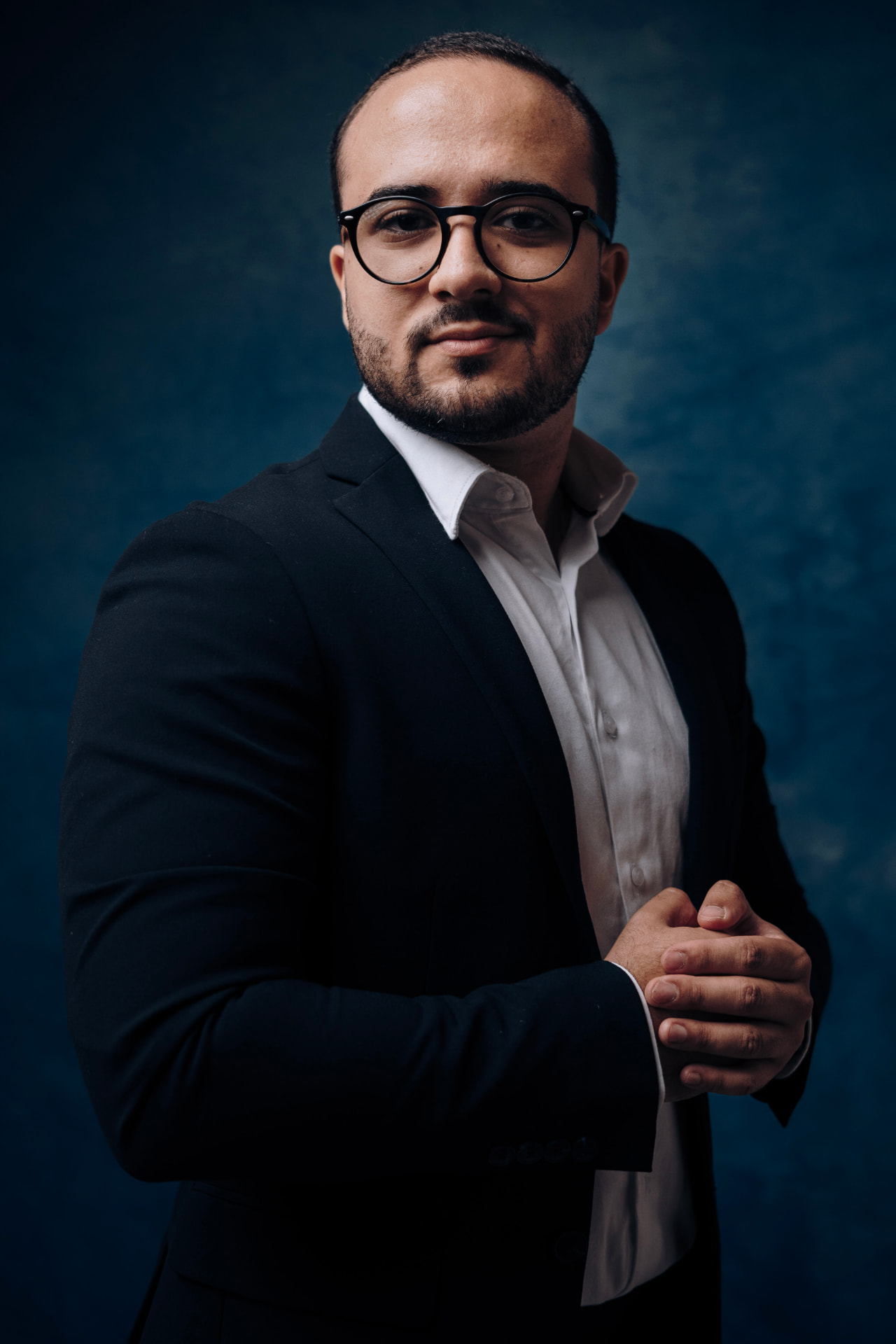 Retrato editorial de homem de blazer escuro com camisa branca e óculos de armação preta, fundo azul-marinho texturizado, fotografado por Lucian Rabello